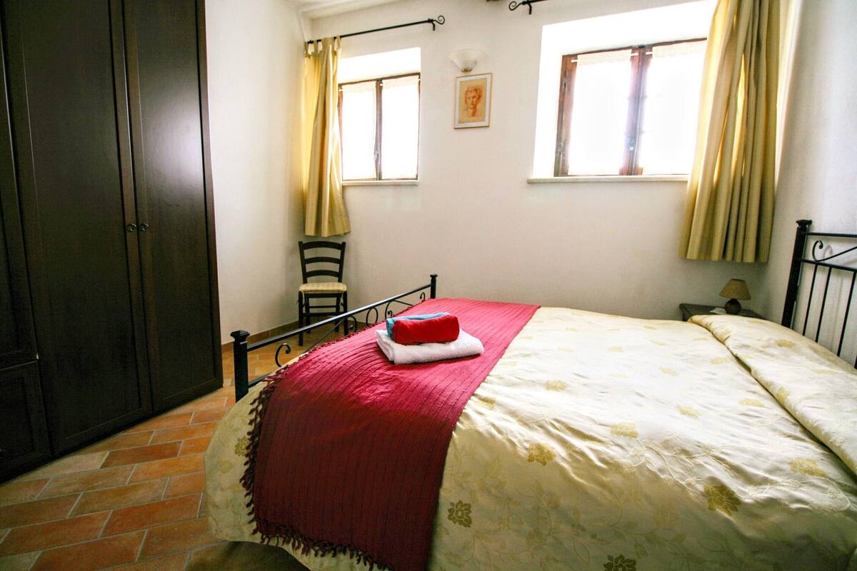 A bedroom with a bed, a chair, a wardrobe, and two windows with curtains.