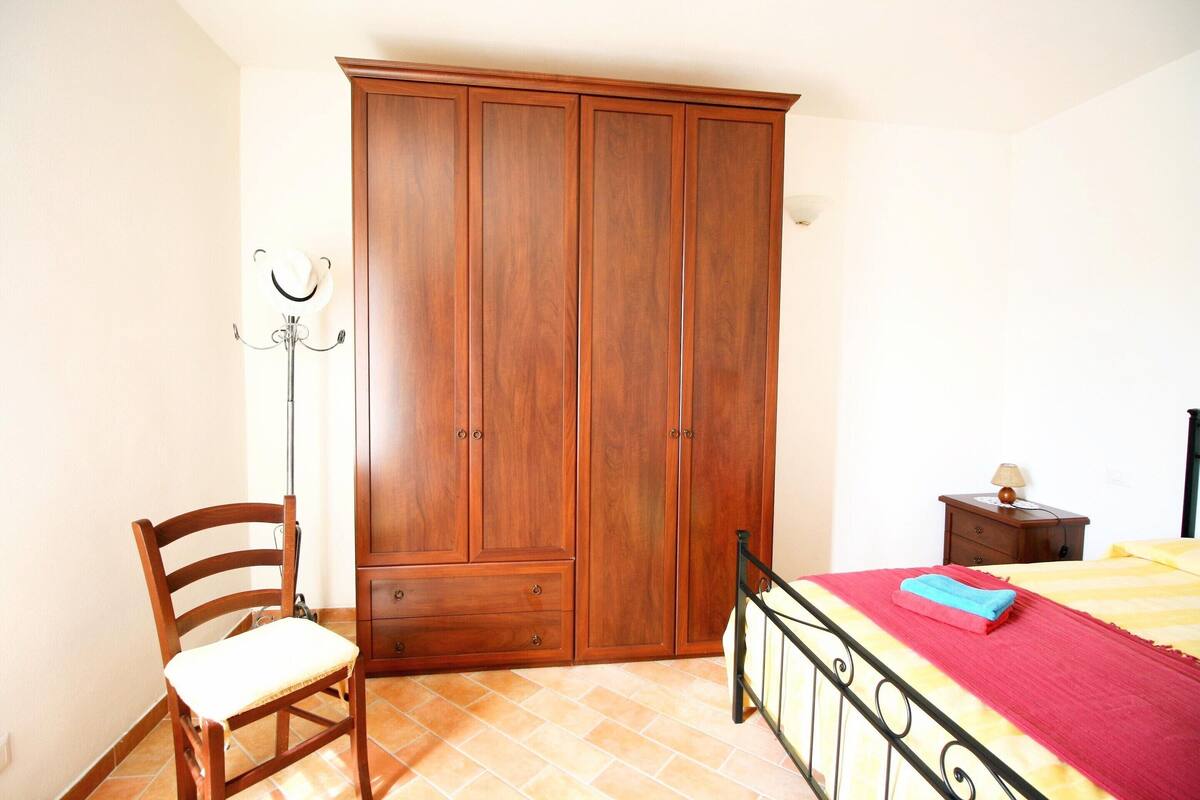 A bedroom with a wooden wardrobe, a chair, a bed with a red and yellow bedspread, and a nightstand with a lamp.