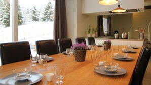Uma sala de jantar com uma mesa de madeira posta para seis pessoas, rodeada por cadeiras pretas. Há plantas em vasos sobre a mesa, e uma área de cozinha é visível ao fundo.