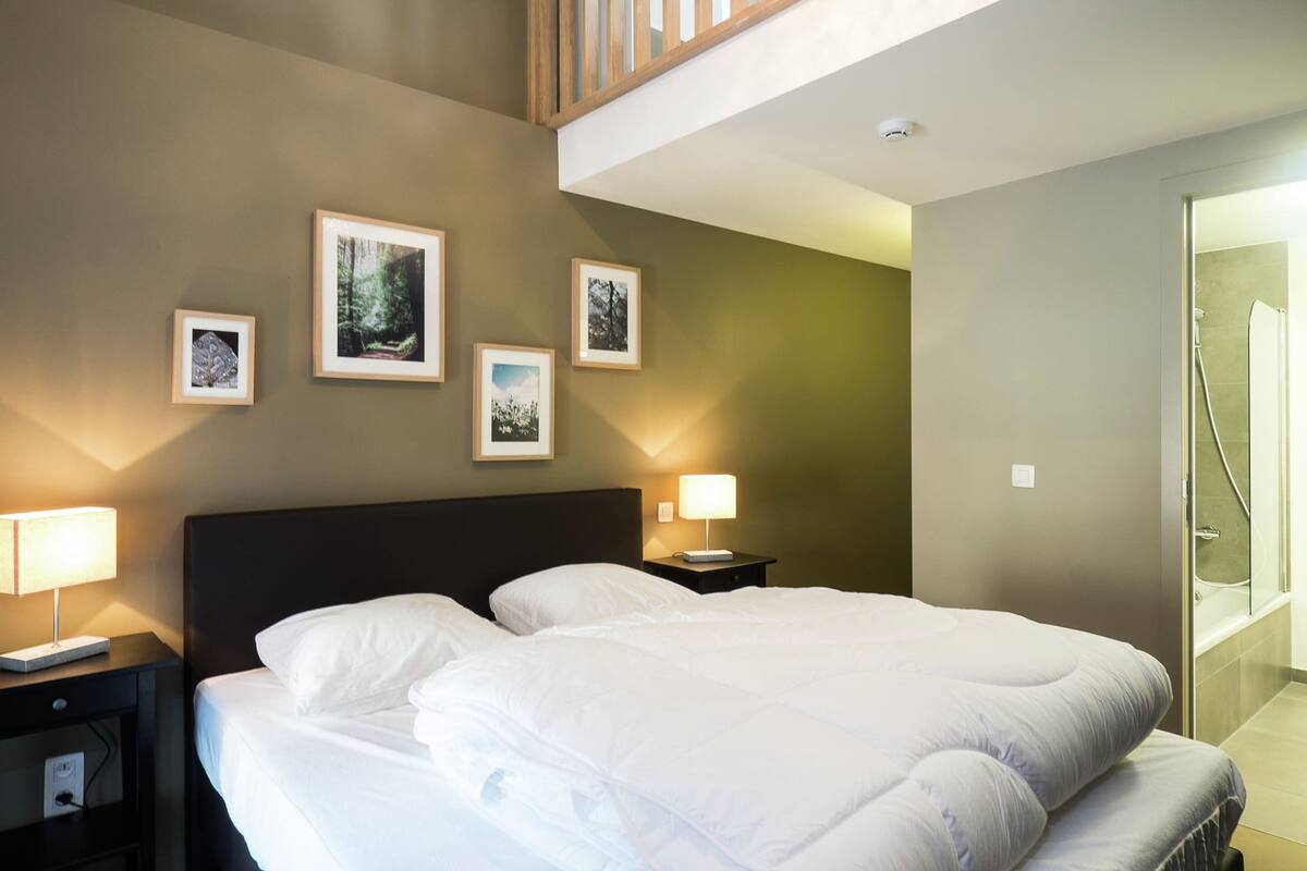 A hotel room with a large bed, two bedside tables, a nightstand, a wall-mounted lamp, and framed pictures.