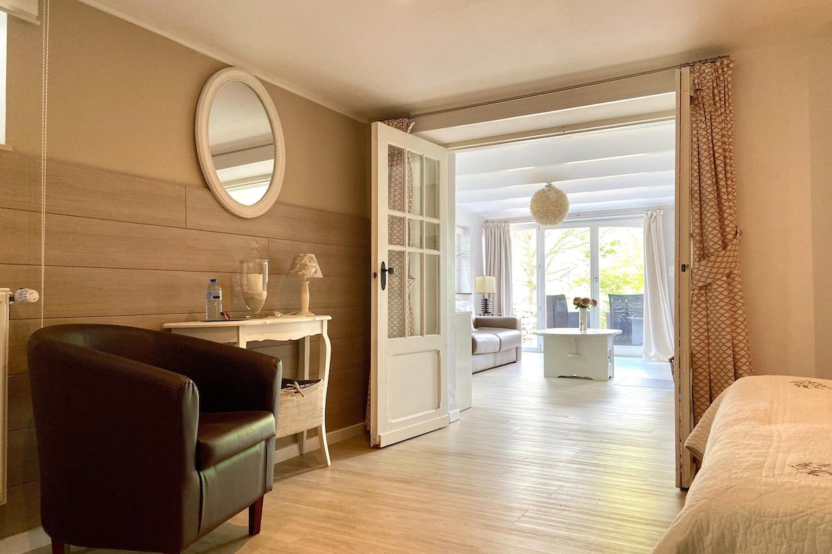 A room with a wooden floor, a bed, a desk with a chair, a mirror, and a door leading to a balcony.