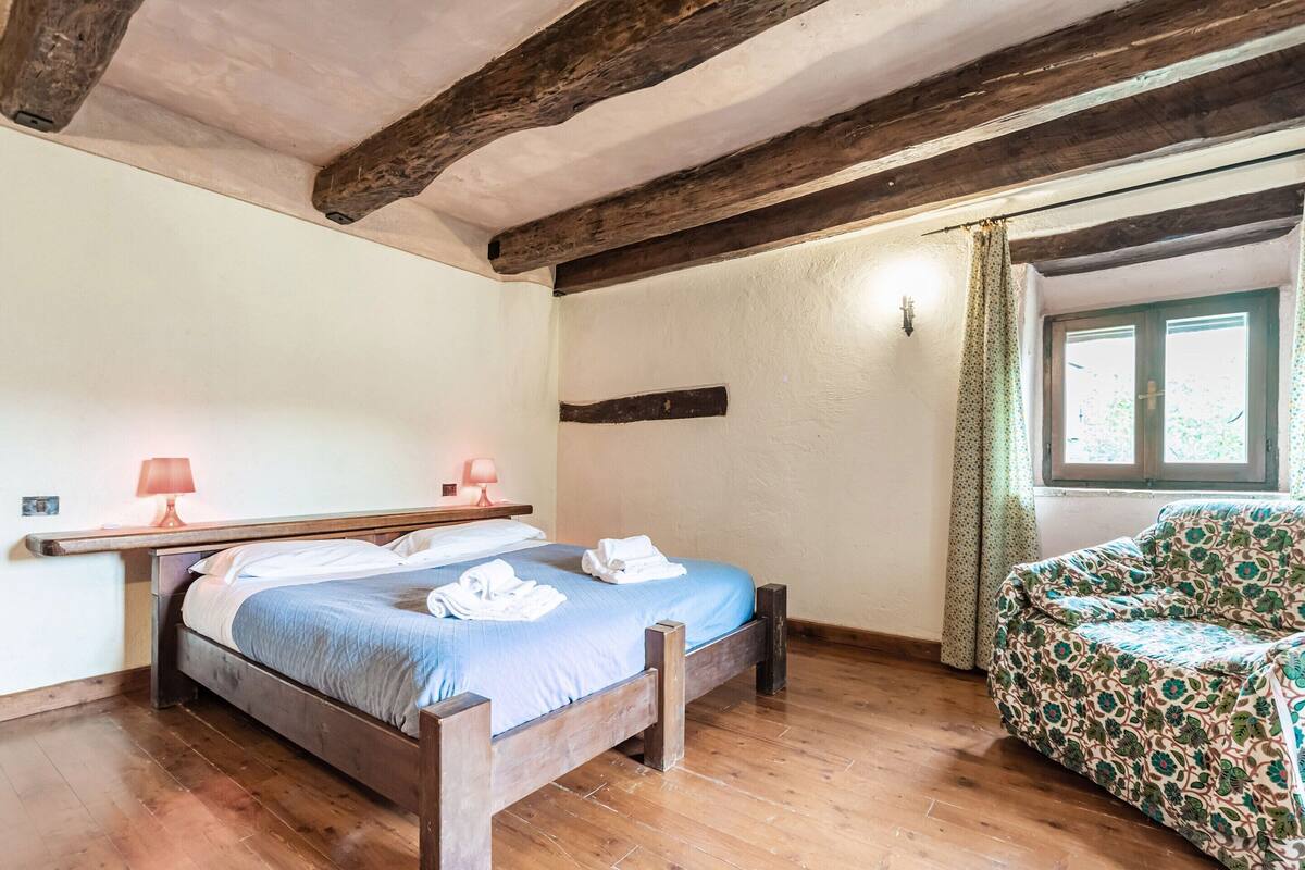 Un dormitorio con una cama de madera, dos lámparas de noche, un sillón estampado con motivos florales y una ventana con cortinas.
