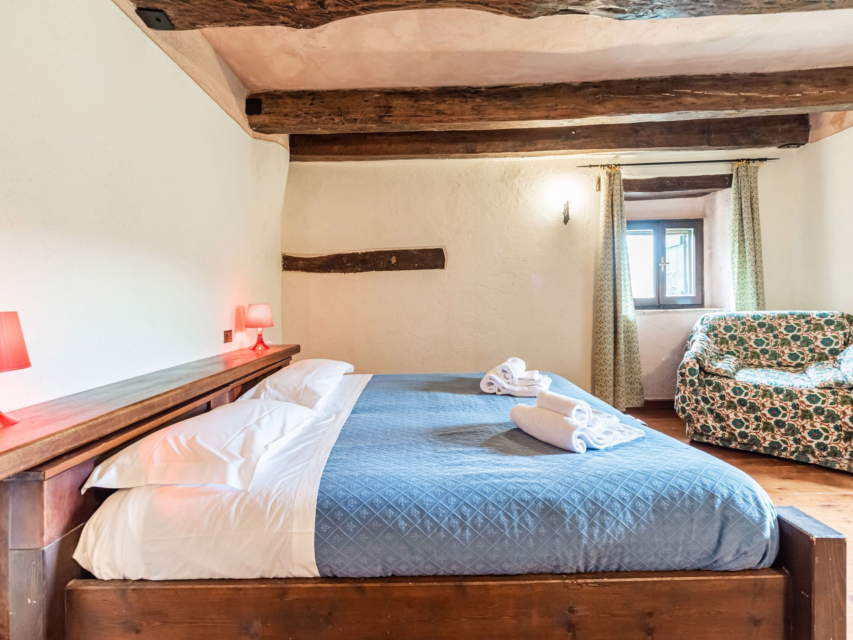 Un dormitorio con una cama de madera, una lámpara roja, un sofá estampado y un ventanal con cortinas.