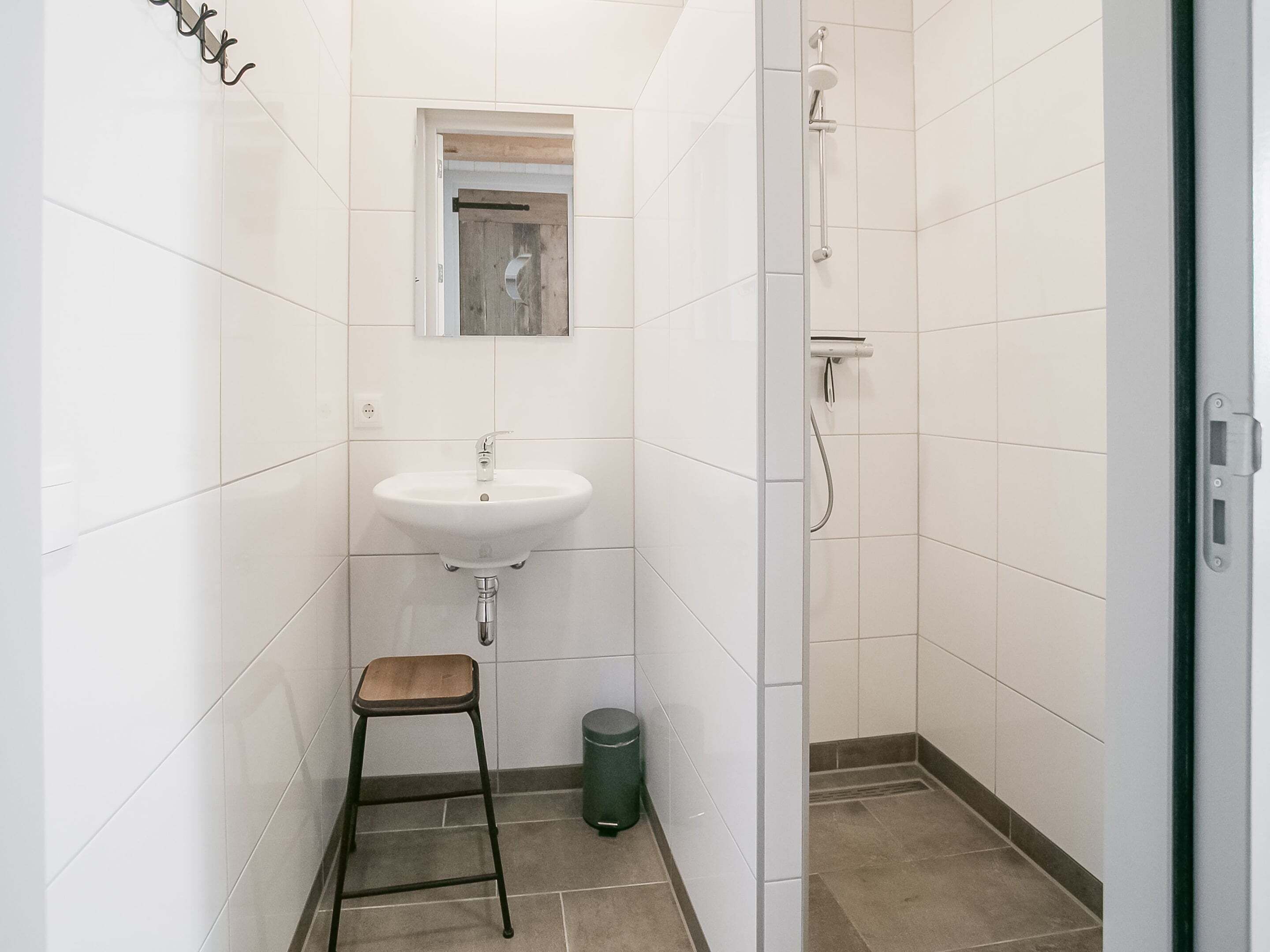 Une petite salle de bain avec un lavabo blanc, un miroir, une douche et un tabouret.