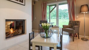 A living room with a fireplace, two armchairs, a dining table with a vase of flowers, and a view of the outdoors through large windows with striped curtains. - Luxury Holiday Home in Thirimont Liege With Private Garden (Waimes)