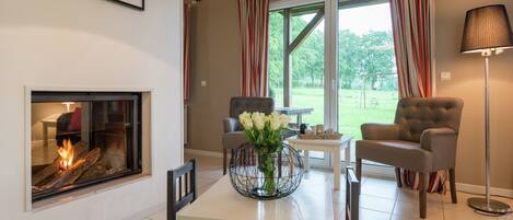 Una sala de estar con chimenea, dos sillones, una mesa de comedor con un jarrón de flores y vistas al exterior a través de amplias ventanas con cortinas a rayas.