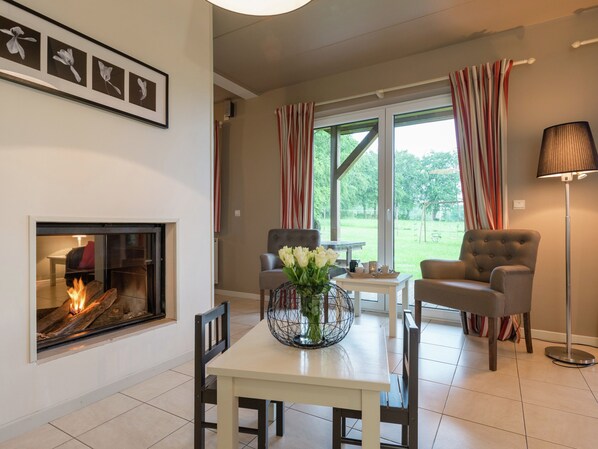 A living room with a fireplace, two armchairs, a dining table with a vase of flowers, and a view of the outdoors through large windows with striped curtains. - Luxury Holiday Home in Thirimont Liege With Private Garden (Waimes)