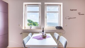 Un coin repas avec une table dressée pour deux personnes, une bouteille de vin et un panier de pain. Deux fenêtres offrent une vue sur la verdure et la mer.