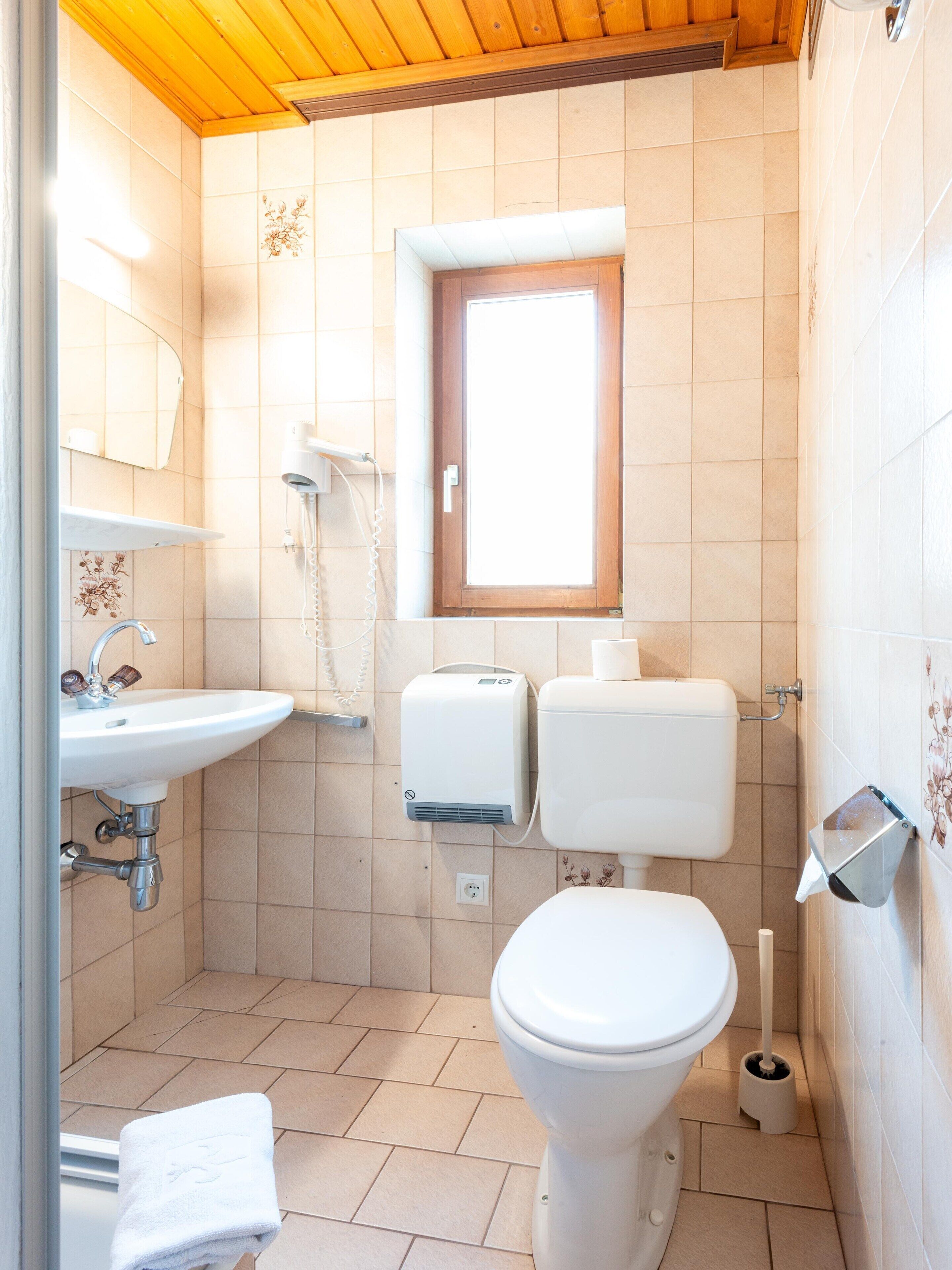 A small bathroom with a sink, toilet, and shower. There is a window above the toilet and a towel hanging on the wall.