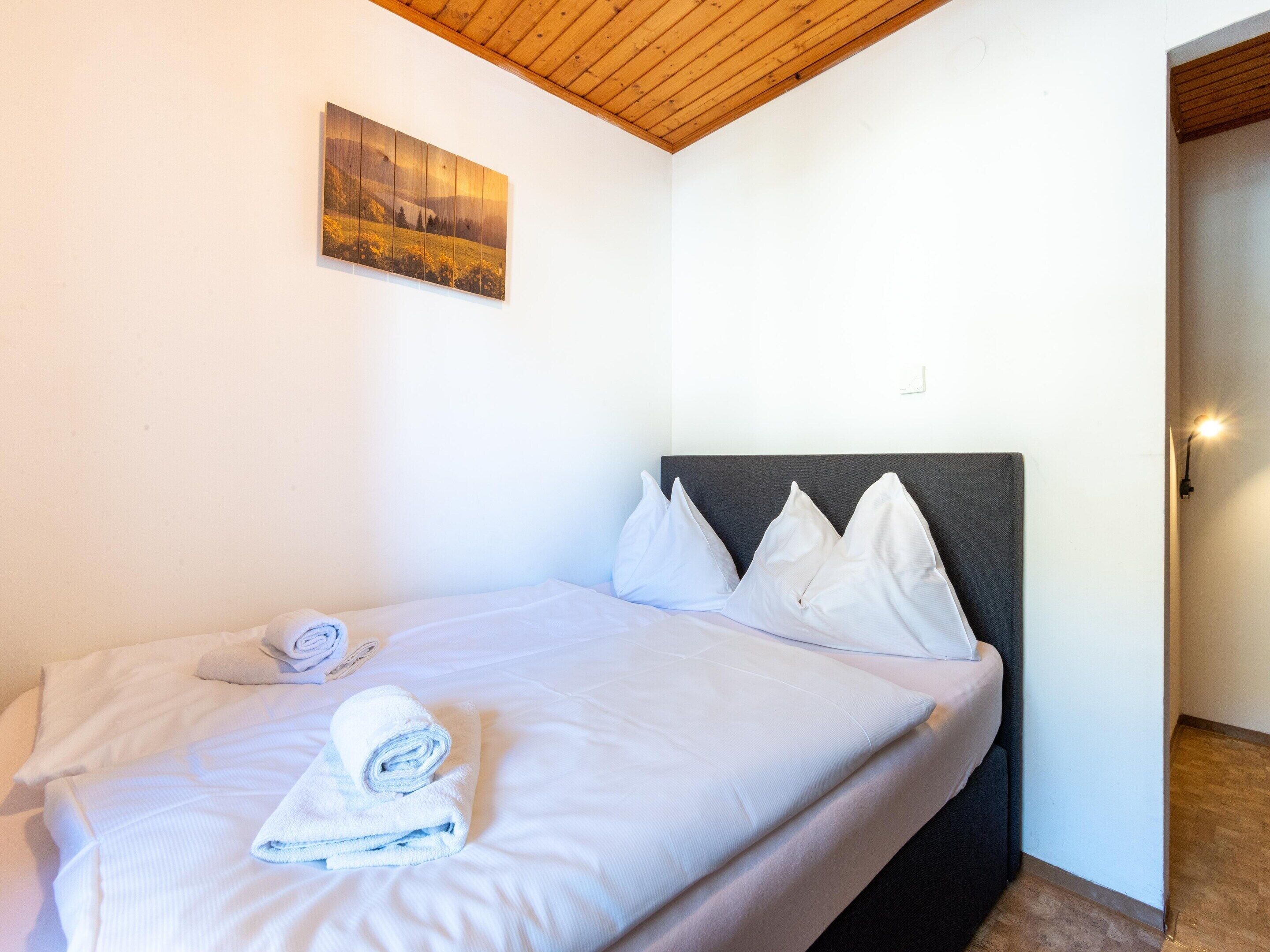 Een net opgemaakt bed met witte lakens en kussens, een houten plafond en een landschapsschilderij aan de muur.