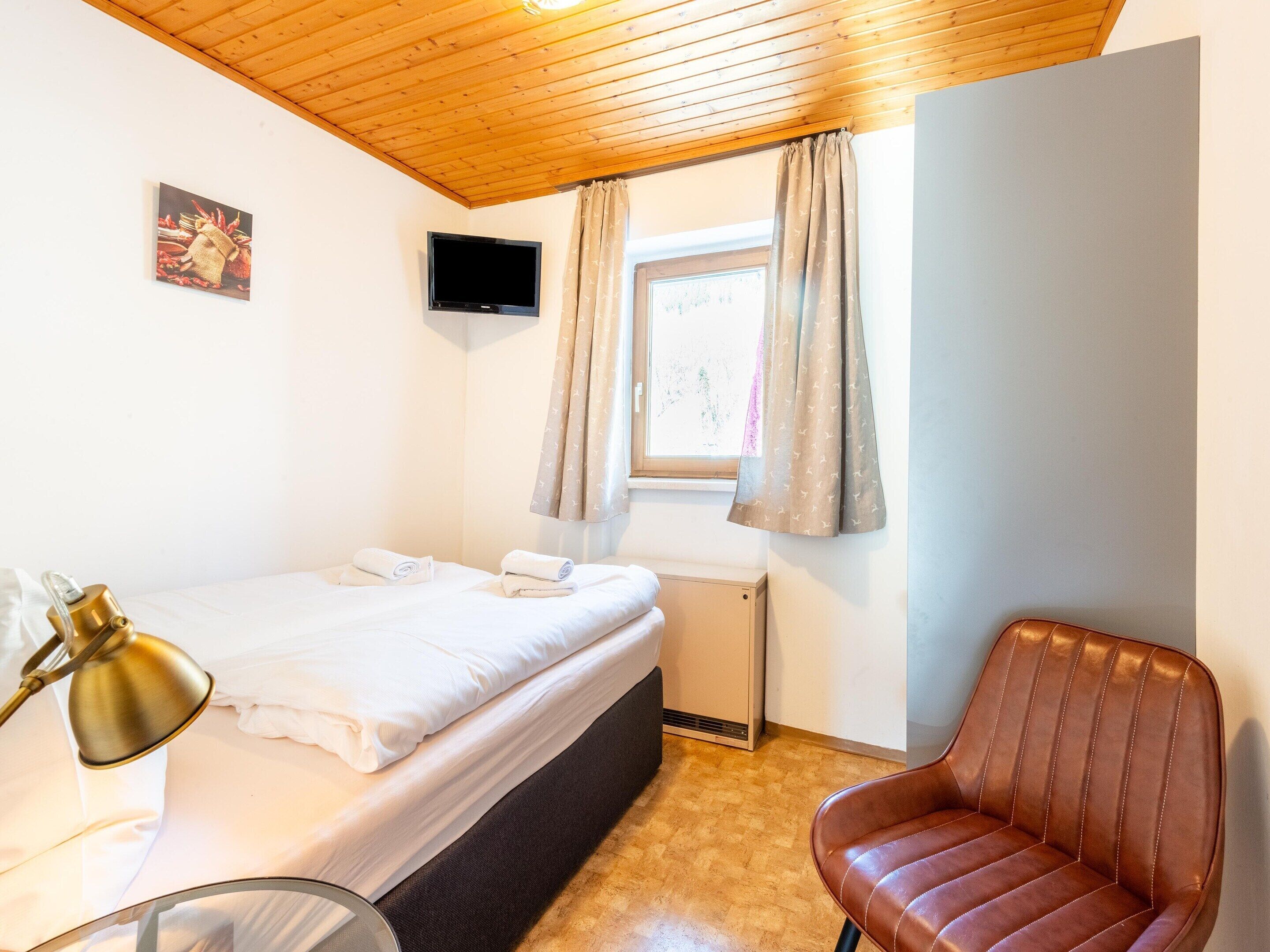A bedroom with a bed, a chair, a TV, and a window with curtains.