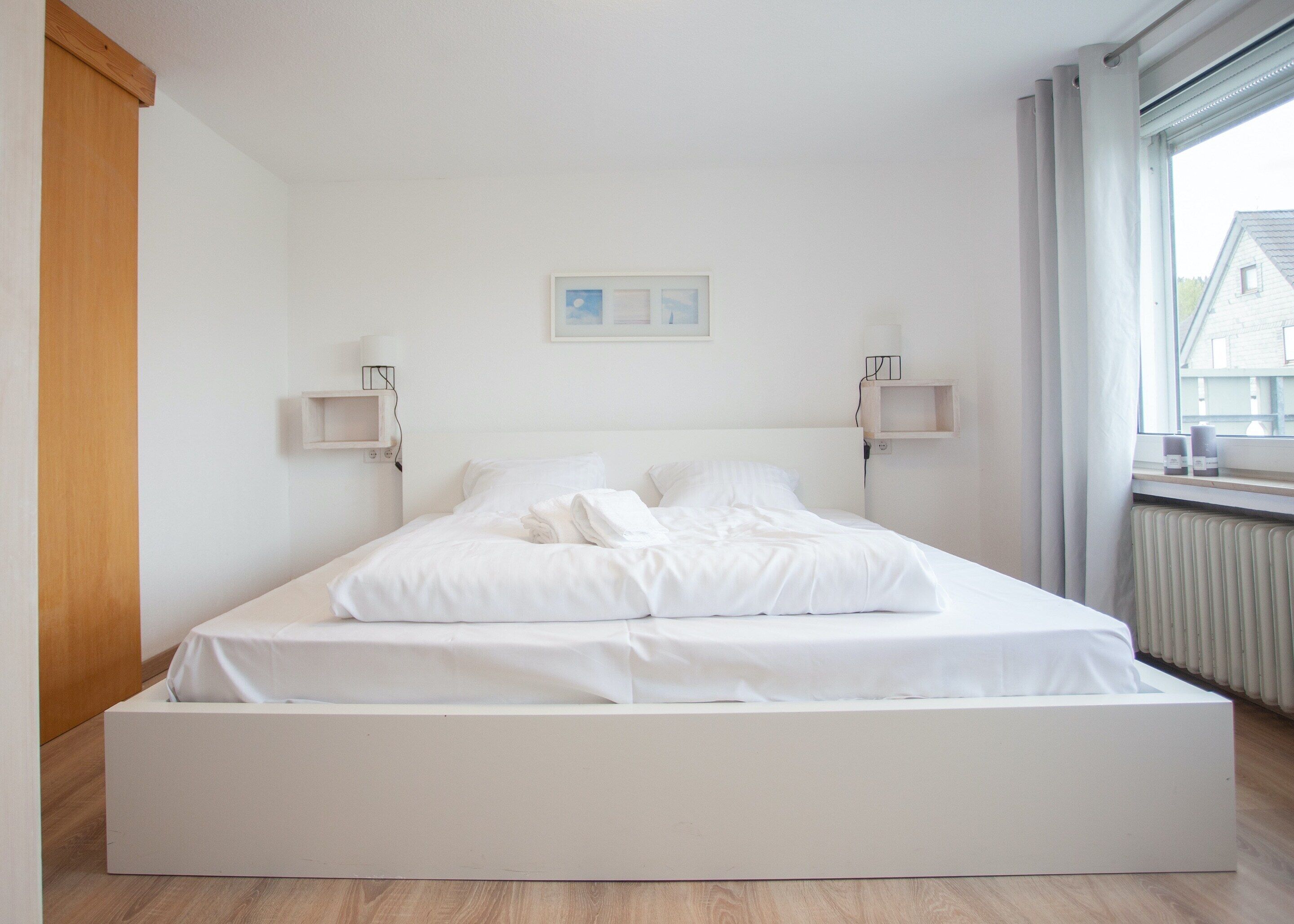 A spacious bedroom with a large bed, wooden floor, white walls, and a window with a view of buildings.