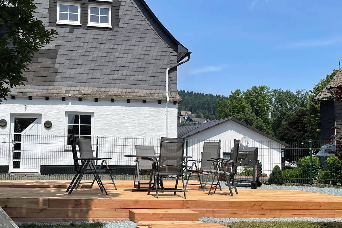 Une maison à l’extérieur blanc, avec un toit gris et une terrasse en bois aménagée avec du mobilier d’extérieur.