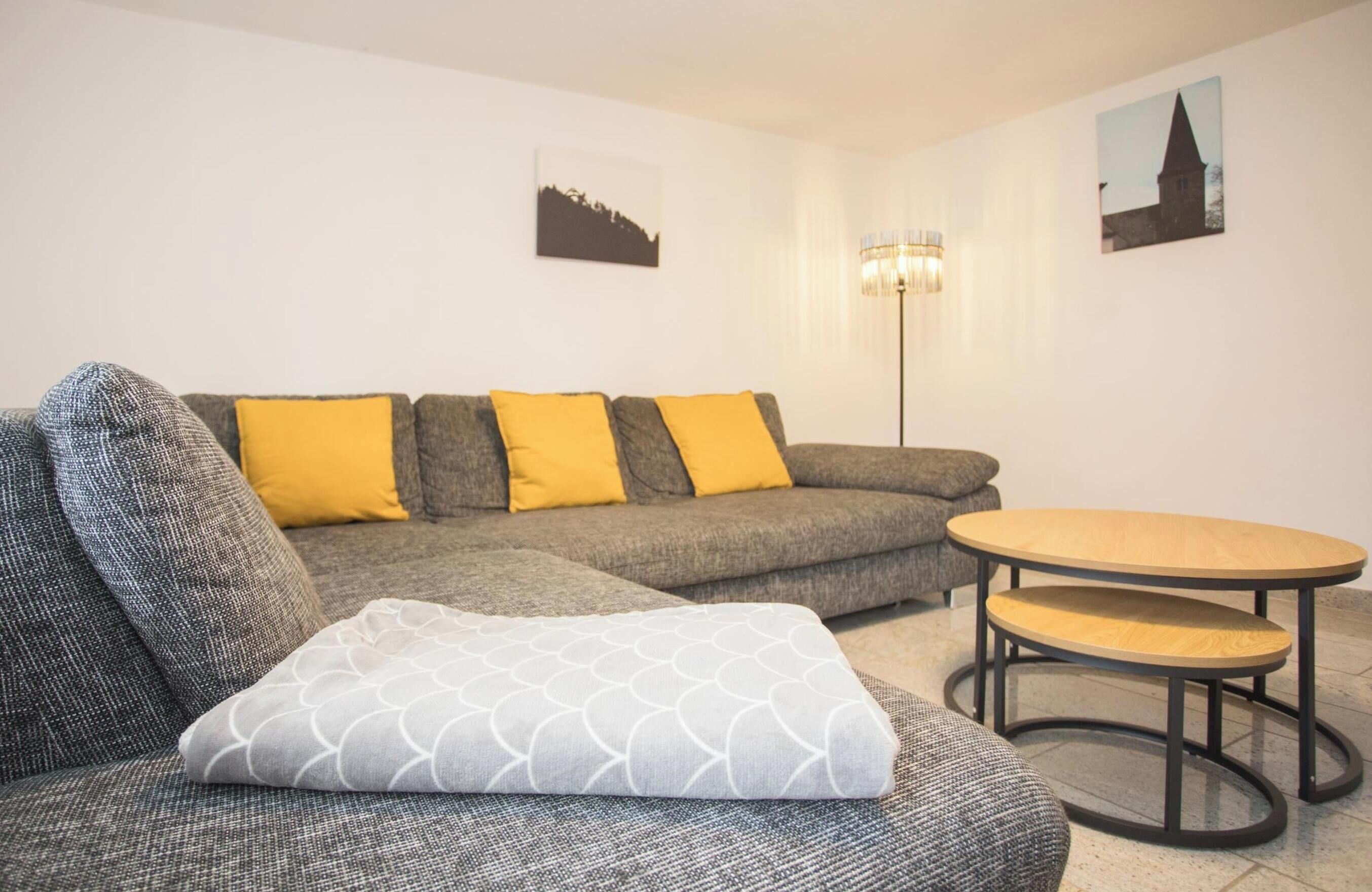 Ein modernes Wohnzimmer mit einem grauen Ecksofa, gelben Kissen, einem runden Couchtisch aus Holz und zwei an der Wand hängenden Bildern.