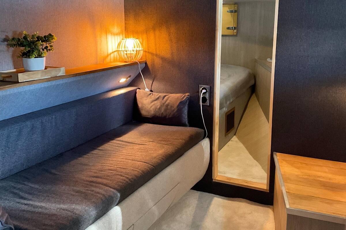 A compact, modern room with a built-in sofa, a bedside table, a lamp, and a mirror.