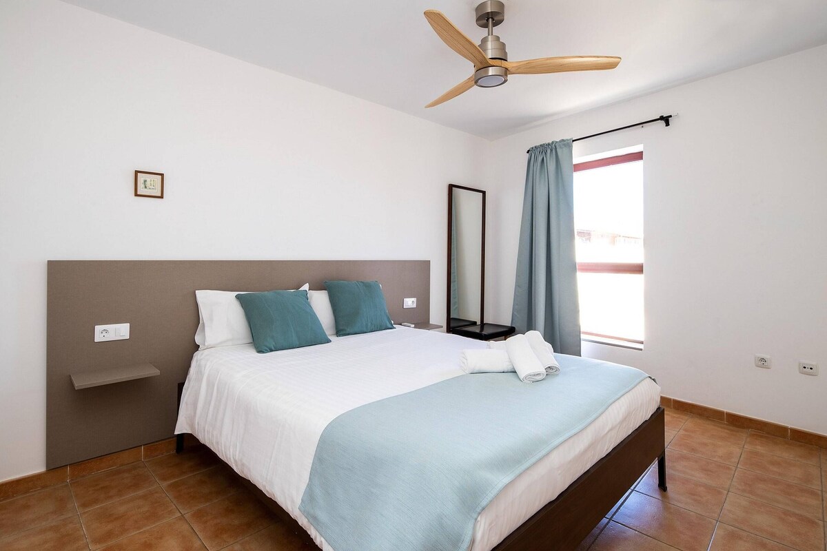 Un dormitorio con cama, ventilador de techo, ventana con cortinas y mesita de noche.