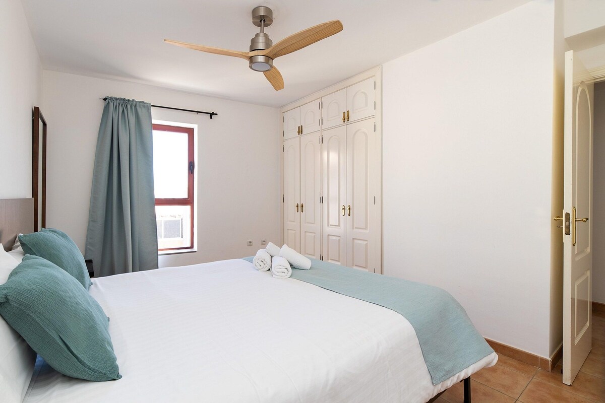 Un dormitorio con cama, ventilador de techo, armario y ventana con cortinas.