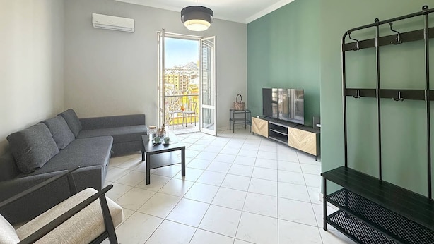 Un salon moderne avec un canapé d’angle, une table basse, un téléviseur et un balcon offrant une vue sur les immeubles.