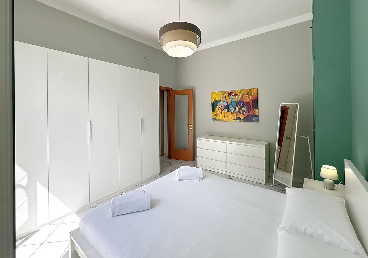 Een moderne slaapkamer met een groot bed, een kledingkast, een dressoir, een spiegel en een schilderij aan de muur.