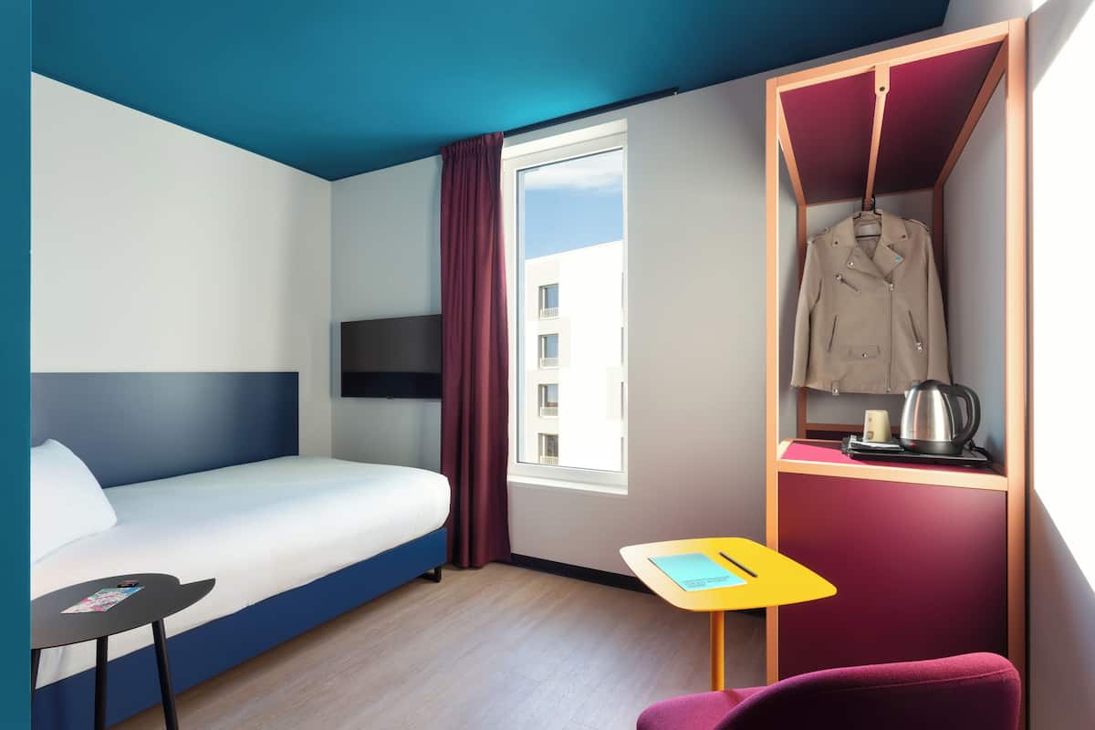 A hotel room with a bed, a desk, a chair, a coat rack with a jacket, a window, and a view of buildings.