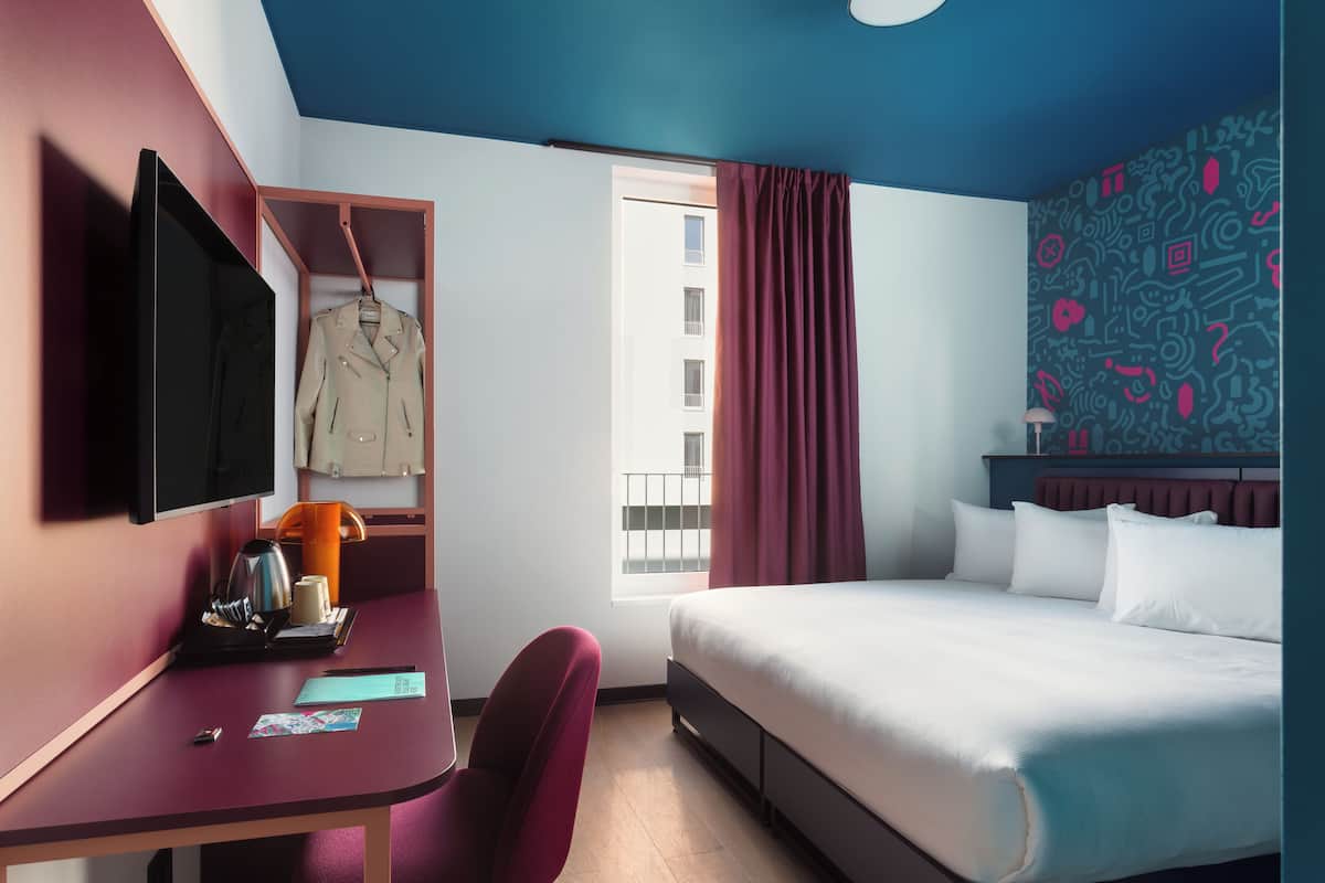 A hotel room with a bed, a desk, a chair, a TV, and a window with curtains.