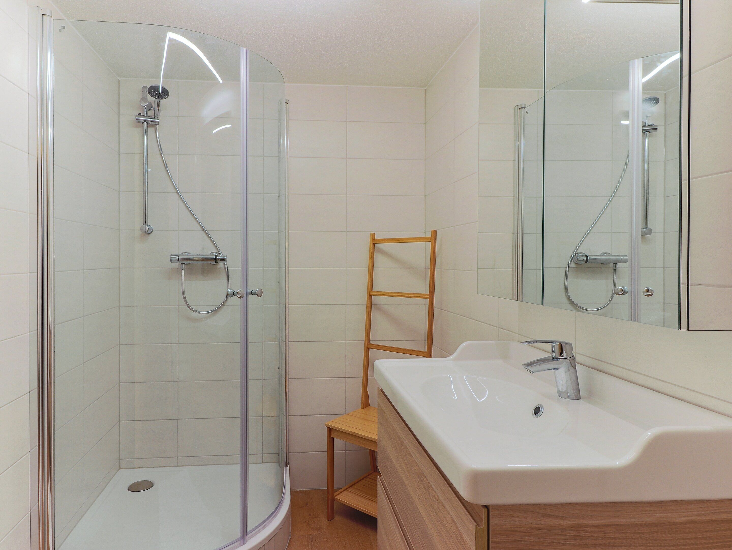 A modern bathroom with a glass shower enclosure, a wooden chair, and a white sink with a mirror above it.