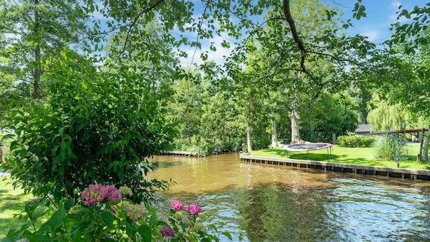A tranquil riverside garden with a wooden dock, lush greenery, and pink hydrangea flowers. - Attractive Farmhouse in Giethoorn With Garden (Giethoorn)