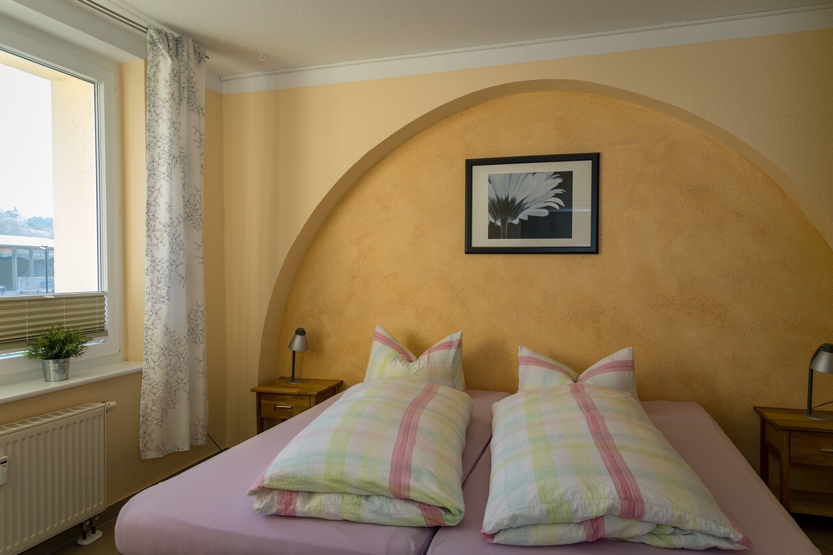 A bedroom with a bed, two bedside tables, a window with a curtain, a radiator, and a framed picture on the wall.