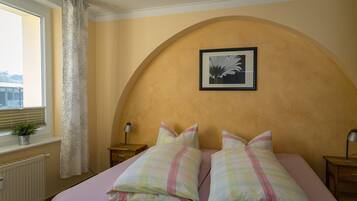 A bedroom with a bed, two bedside tables, a window with a curtain, a radiator, and a framed picture on the wall.