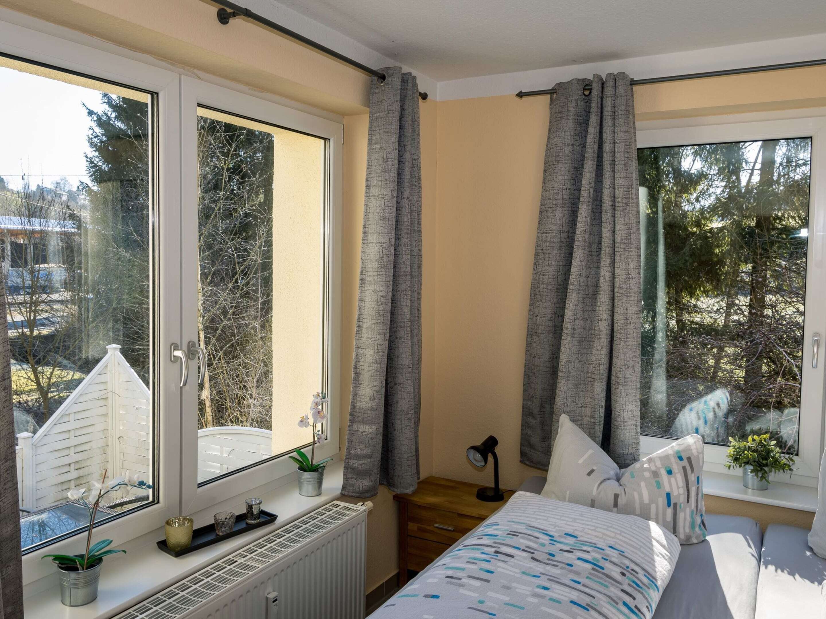 A bedroom with a large window, a radiator, a bed, a nightstand, and a potted plant.