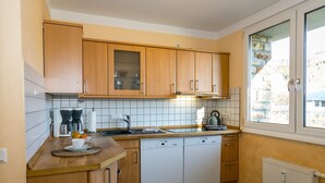 Egy praktikus, kisebb méretű konyha, melyben fa szekrények, mosogatógép és egy ablak található, mely természetes fényt biztosít.