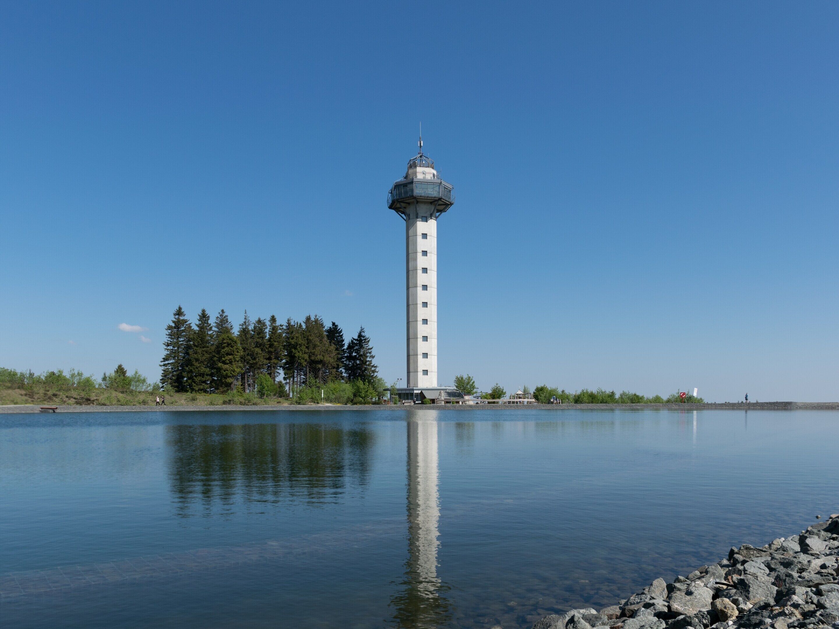 A tall, narrow tower by a calm lake with a clear sky.