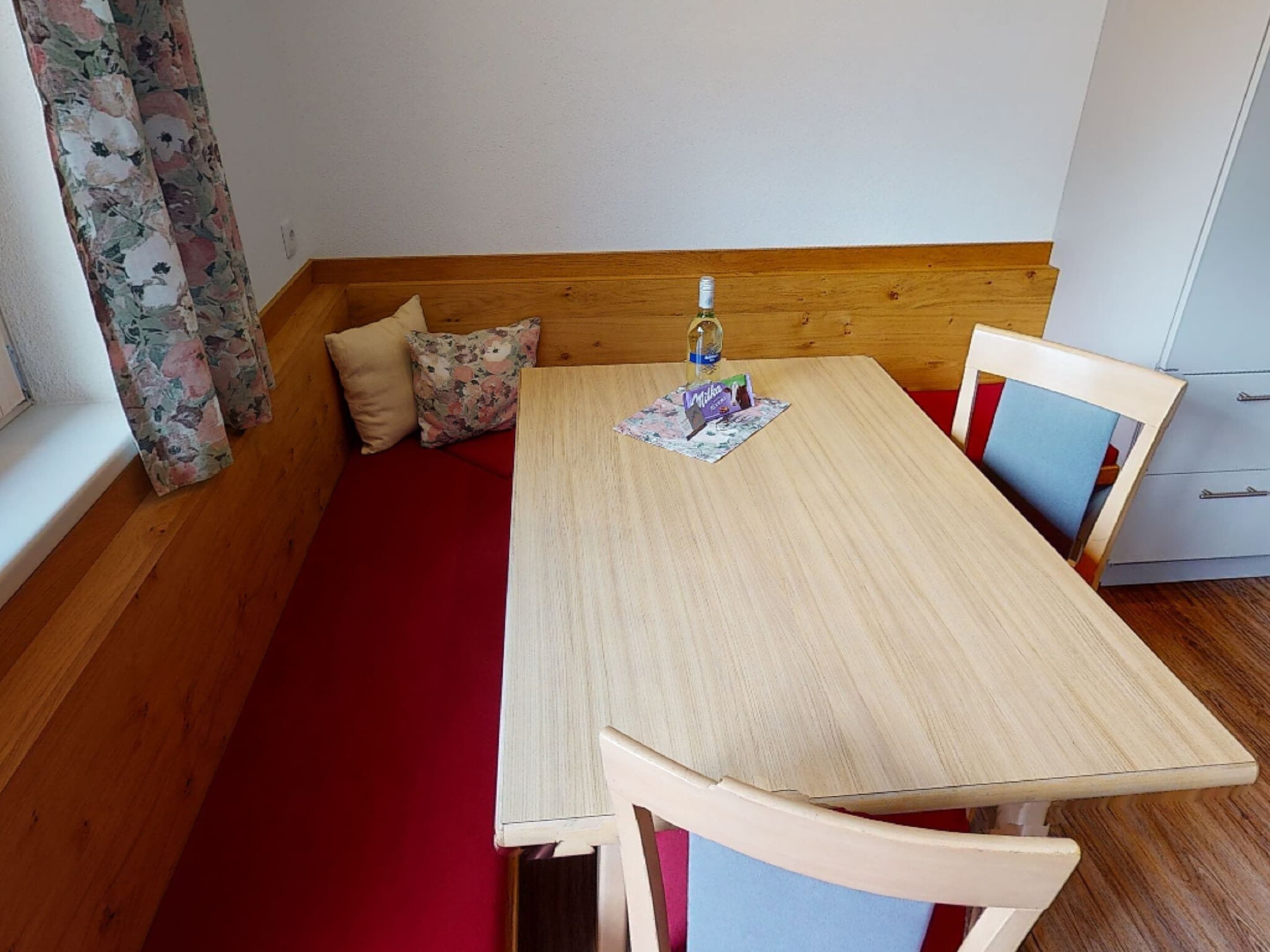 A small, cozy room with a wooden bench, a wooden table, and a chair. There is a window with floral curtains and a white cabinet with drawers.