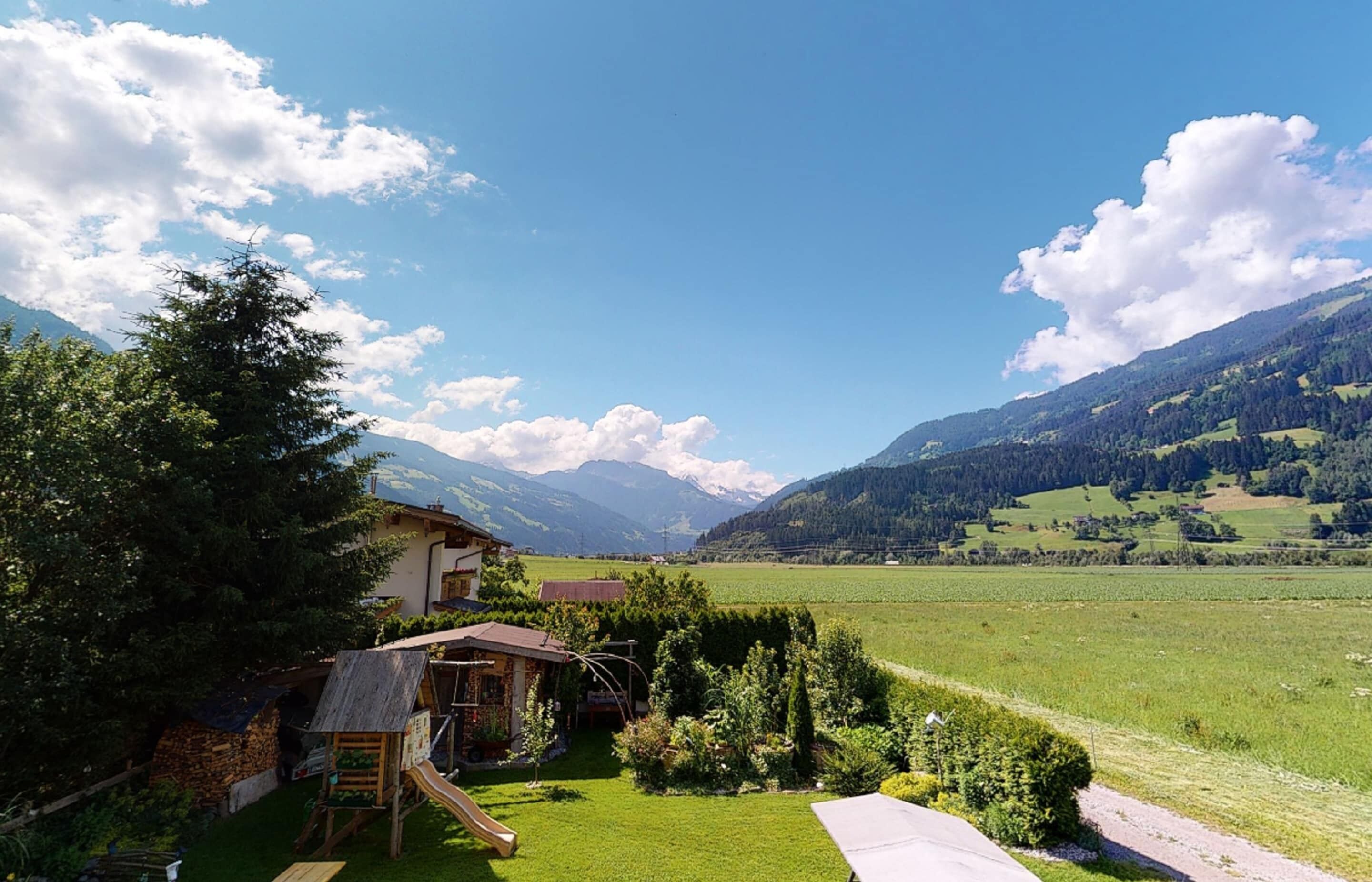 A rural house with a wooden play area, surrounded by a lush green field and mountains in the background.