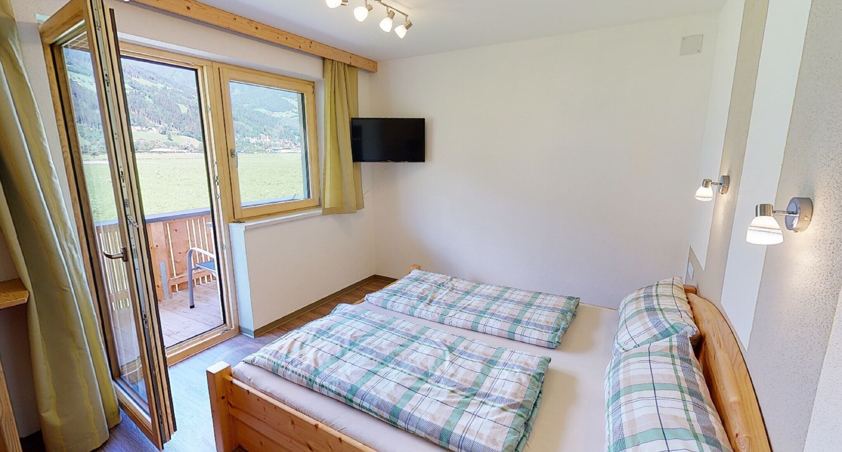 A bedroom with two beds, a TV mounted on the wall, and a large window with curtains.
