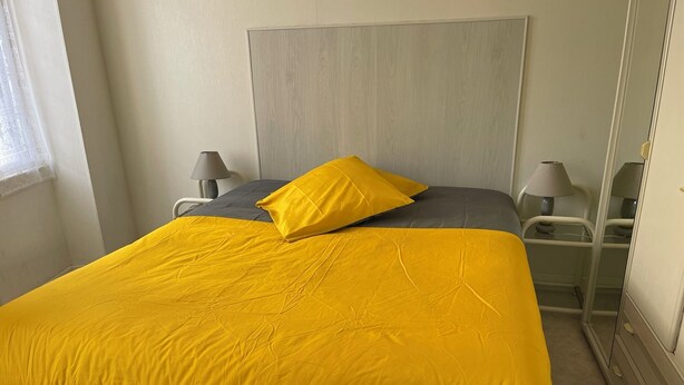 Una cama con cubrecama amarillo y almohada gris, una mesita de noche con lámpara, y un espejo en la pared.