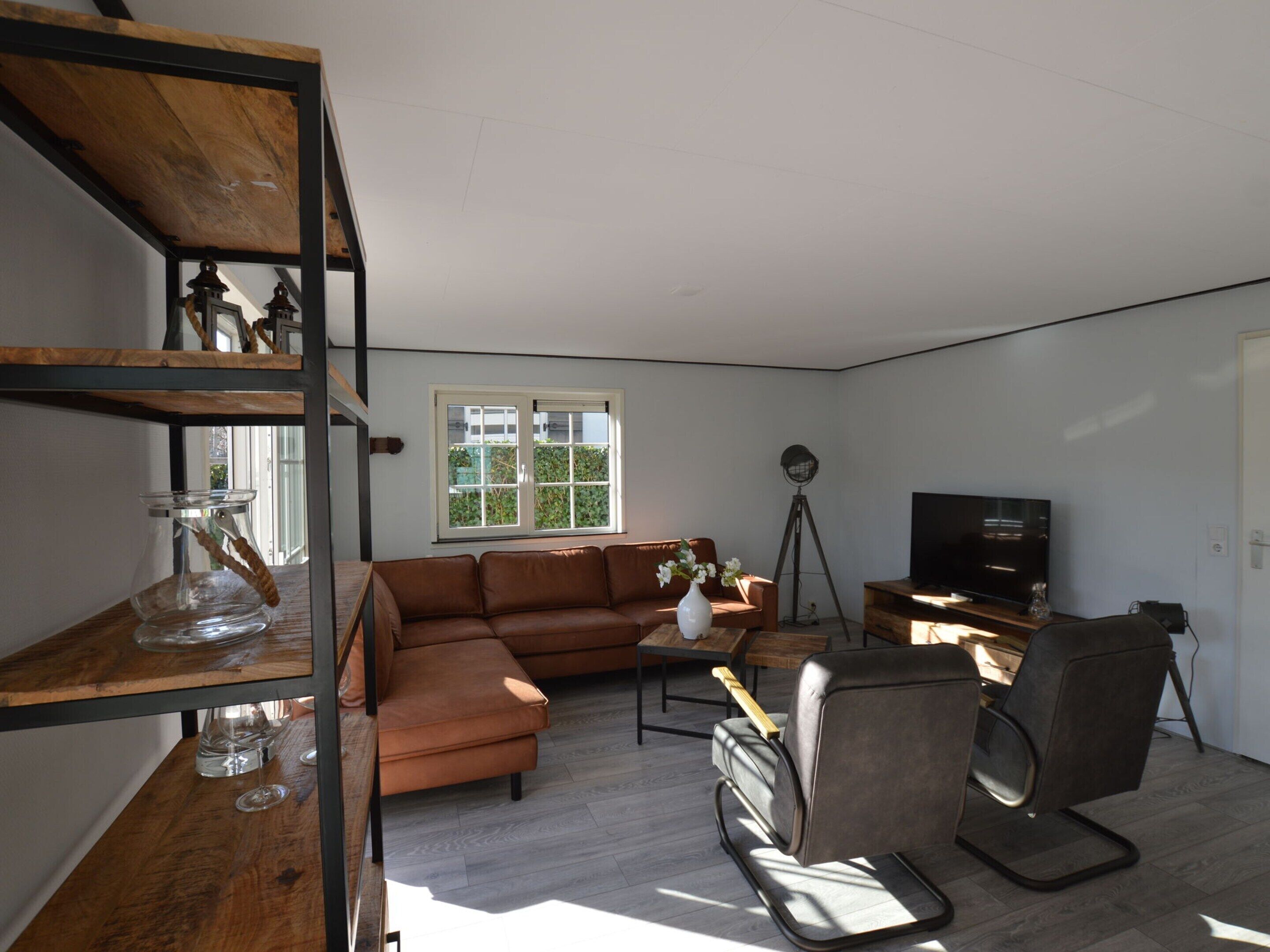 Ein geräumiges Wohnzimmer mit einer braunen Ledersofa, einem Holztisch und einem modernen Sessel. Ein Fernseher ist an der Wand montiert, und eine Stehlampe steht dort.