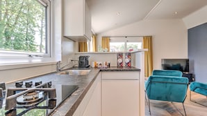A modern kitchen with a gas stove, sink, and a built-in oven. There is a dining area with a teal chair and a flat-screen TV. - Lovely Holiday Home in Kaatsheuvel With Bubble Bath (Kaatsheuvel)