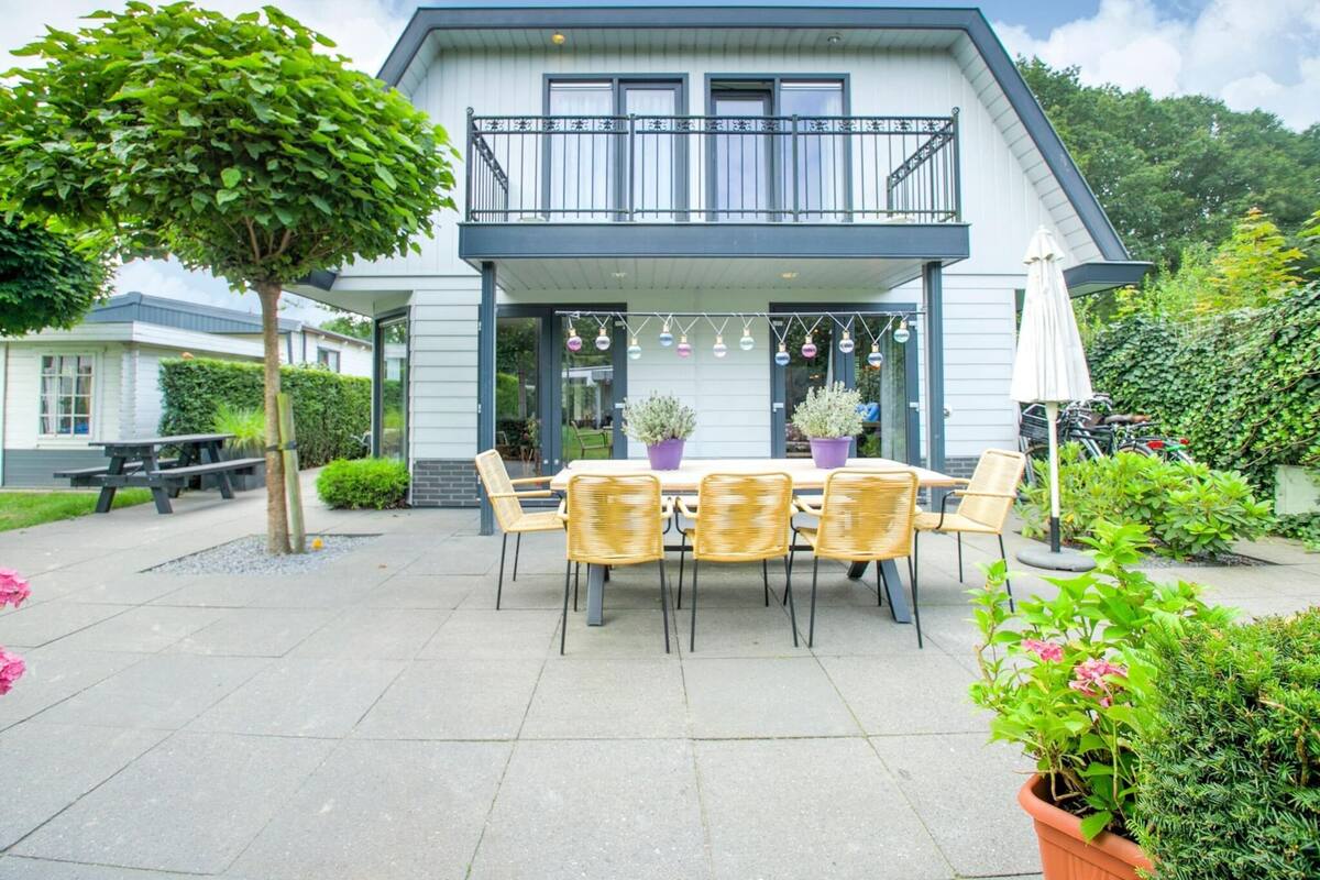 Une maison à deux étages dotée d’un balcon, d’un espace extérieur aménagé avec des chaises et une table en osier, ainsi que de plantes en pot.