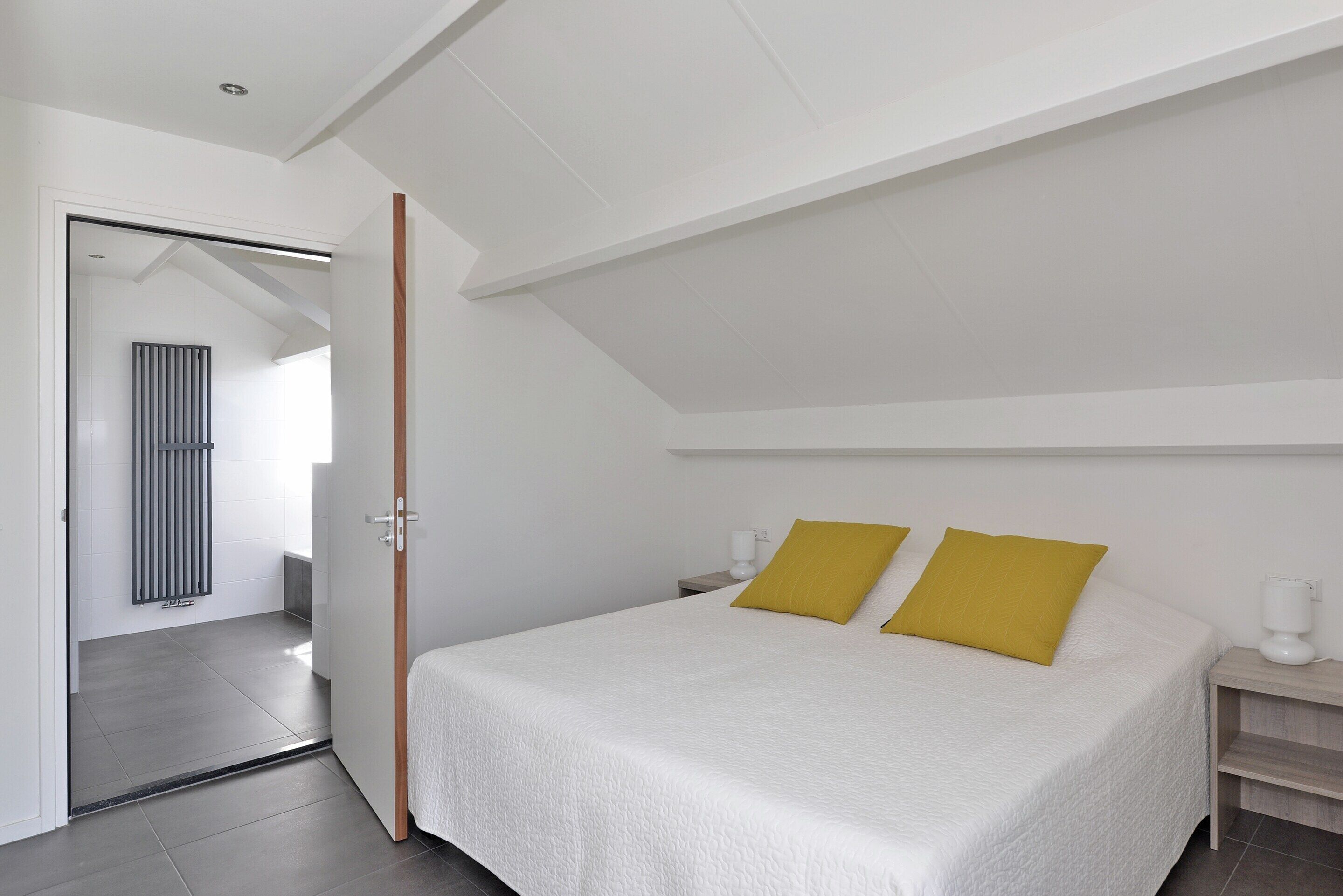 Un dormitorio con cama, dos almohadas, una mesita de noche y un radiador. Se ve una puerta que da a otra habitación.