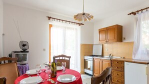 A dining area with a red tablecloth, white plates, and wine glasses. There is a TV, a bottle of wine, and a chair. The kitchen has wooden cabinets and a white refrigerator.