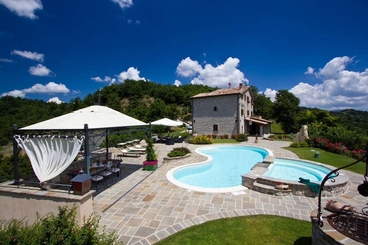A swimming pool with a stone deck, outdoor seating, and a stone building in the background.