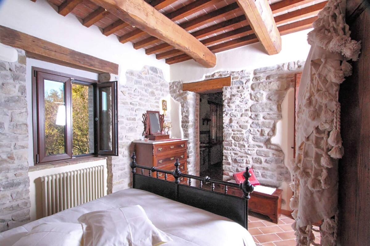 Ein Schlafzimmer mit Steinwand, Holzbalkendecke, einem Bett mit weißen Leinen, einem Schminktisch und einem Spiegel.