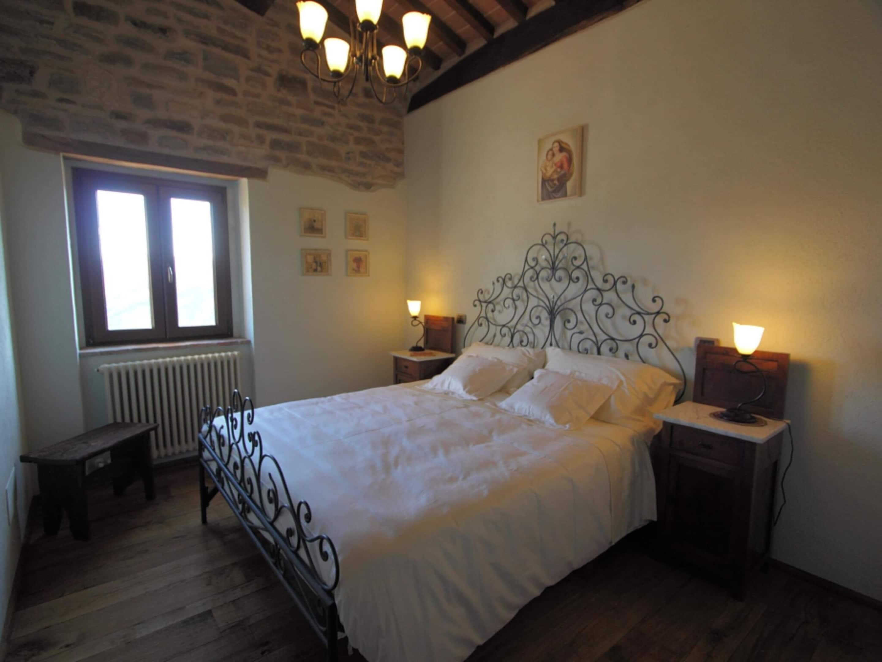 A bedroom with a large bed, a wooden floor, a stone wall, and a window with blinds.