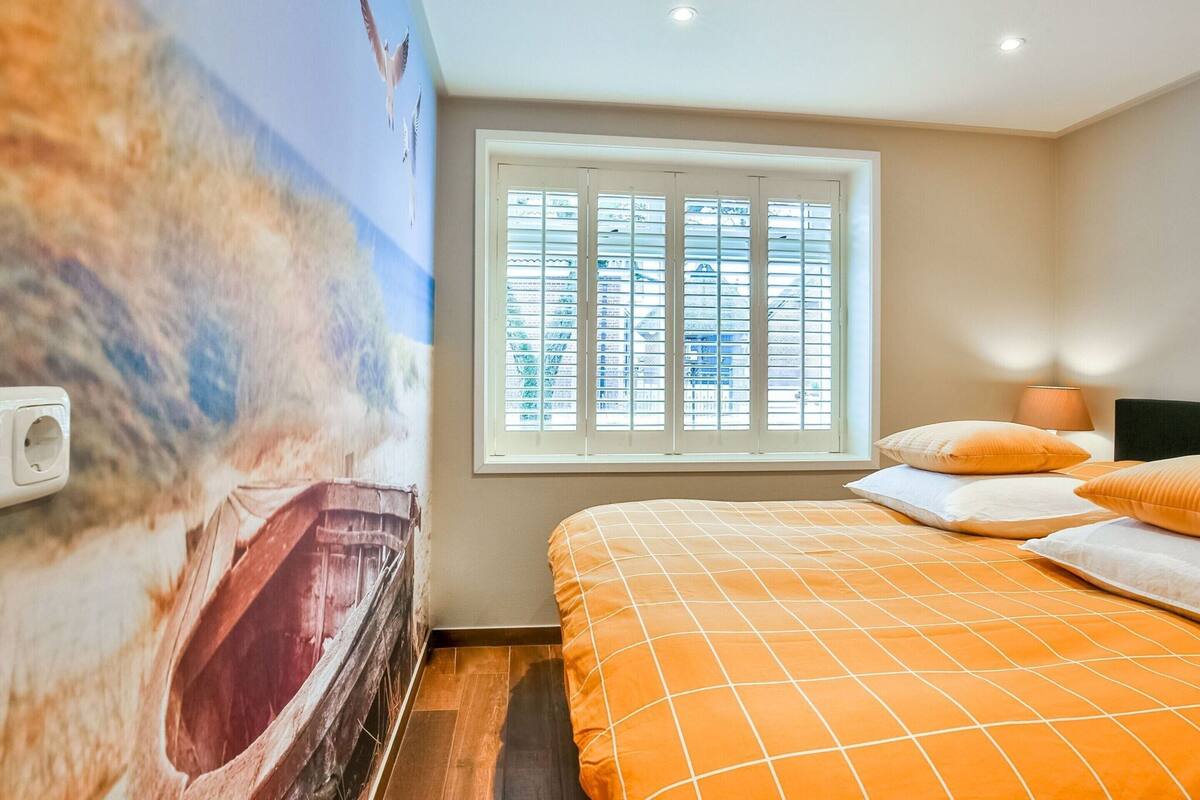 Une chambre avec une grande fresque murale représentant une scène de plage, une fenêtre avec des stores, un lit avec une literie orange et une lampe de chevet.