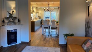 Un salon moderne avec une cheminée, un coin repas avec des chaises en bois et une cuisine visible à travers une porte ouverte.