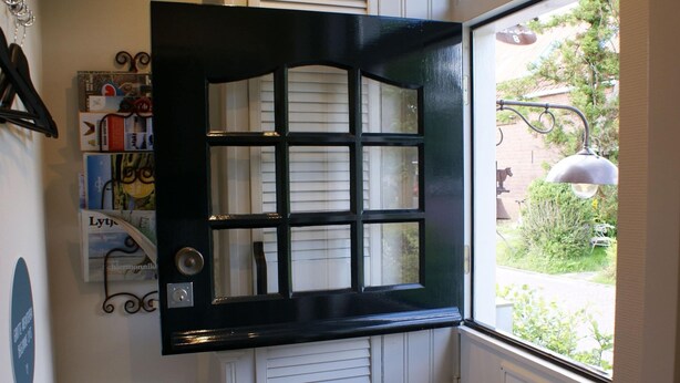 Une porte à cadre noir ornée d’un motif en croisillons, un luminaire fixé au mur et, par la fenêtre, une vue sur un jardin.