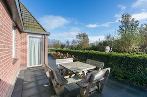 Une terrasse avec une table et des bancs, un bâtiment en briques et une haie.
