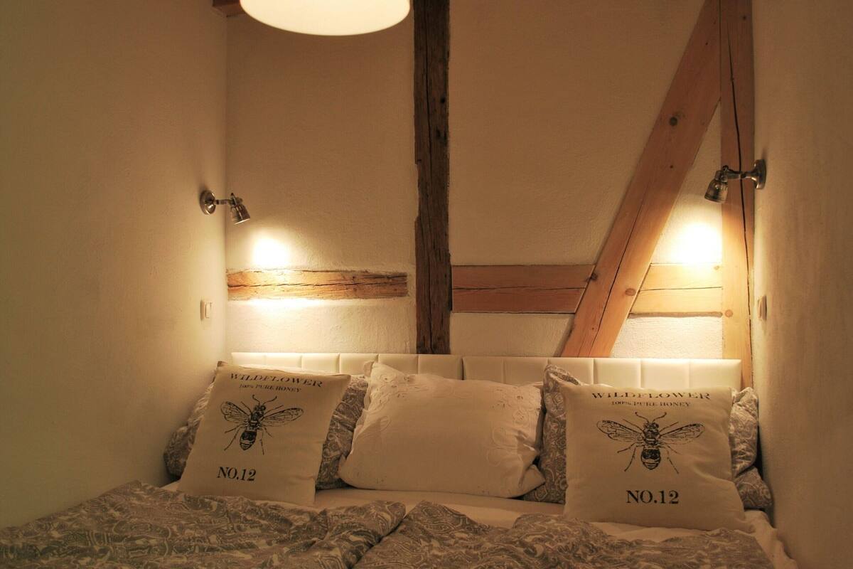 Une chambre avec un plafond en pente, des poutres en bois et deux lampes fixées au mur. On y trouve deux coussins ornés d’un motif d’abeille et portant l’inscription « WILDFLOWER 100 % MIEL PUR, N° 12 ».