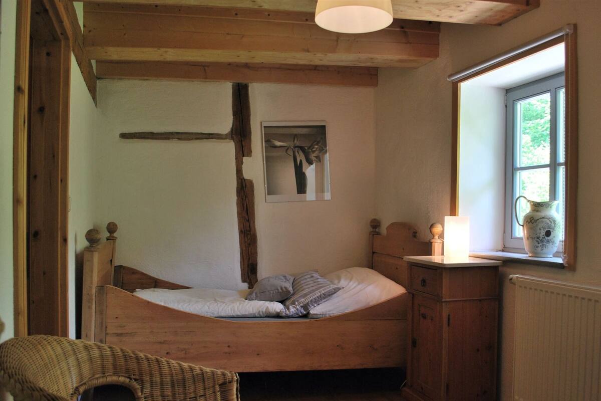 Une pièce aux murs en bois, comprenant un lit, une chaise en osier, une armoire et une fenêtre agrémentée d’un vase.